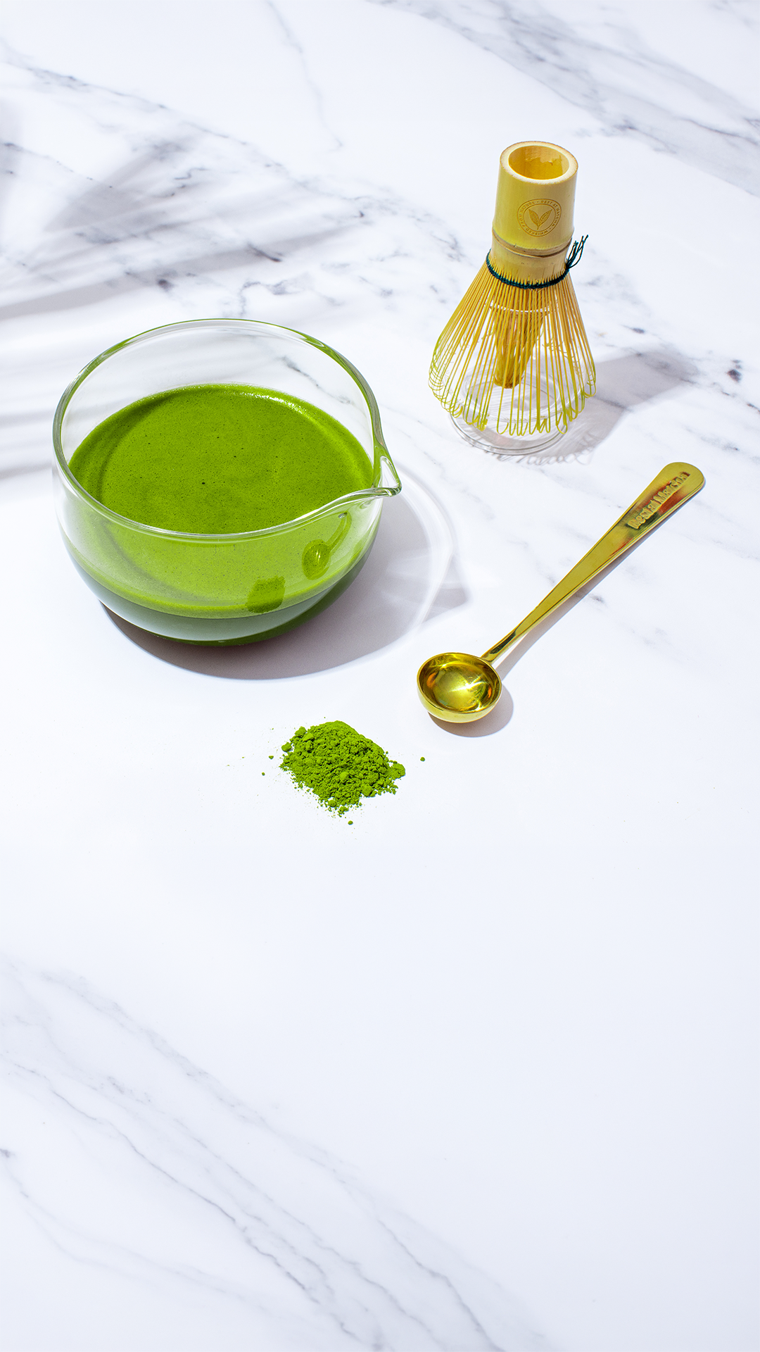 Matcha green tea in a glass bowl with a whisk, scoop, and matcha powder on a white marble surface.