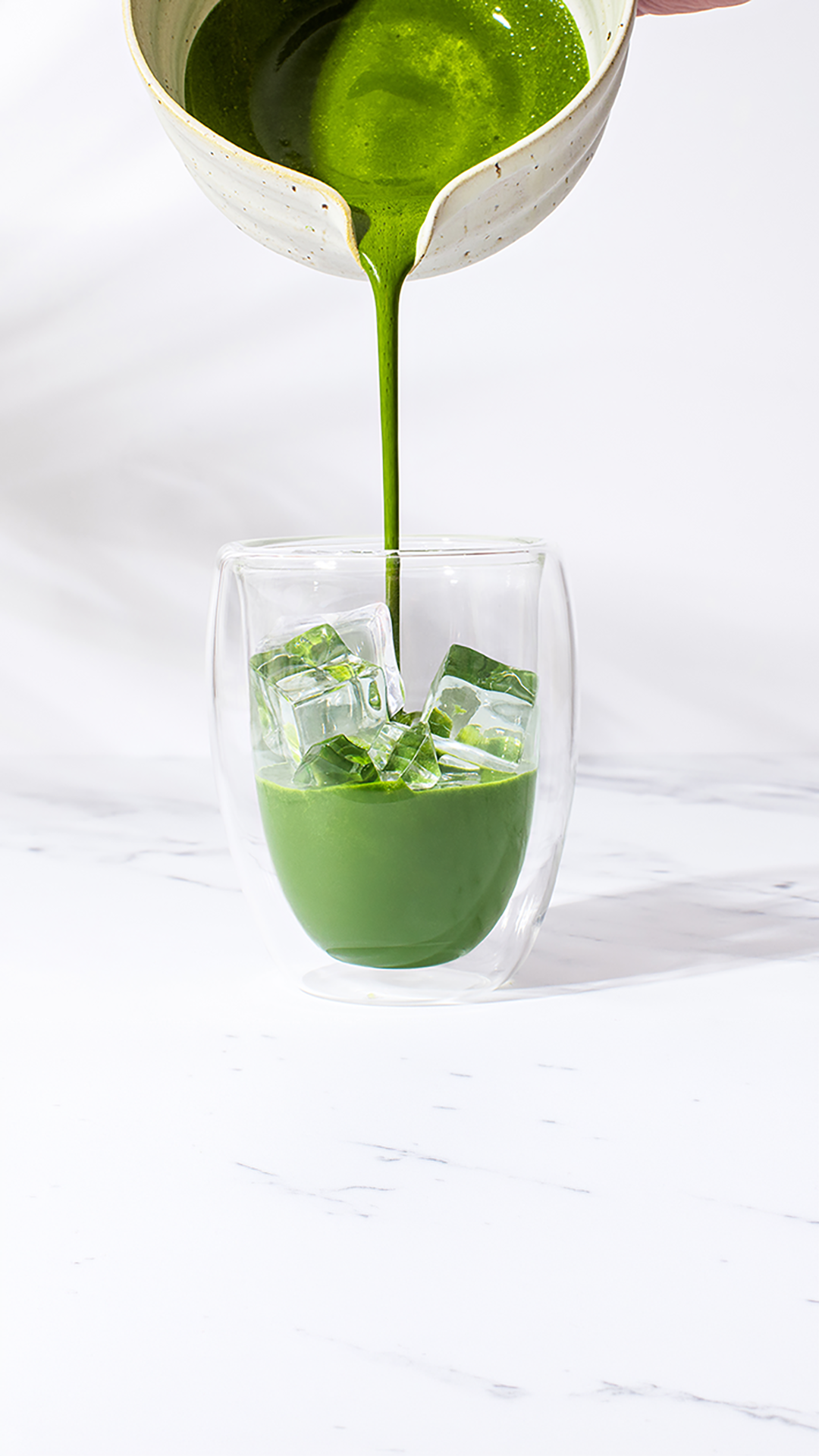 Close up of Ceremonial grade matcha being poured from a ceramic chawan into an iced matcha latte glass on a marble surface