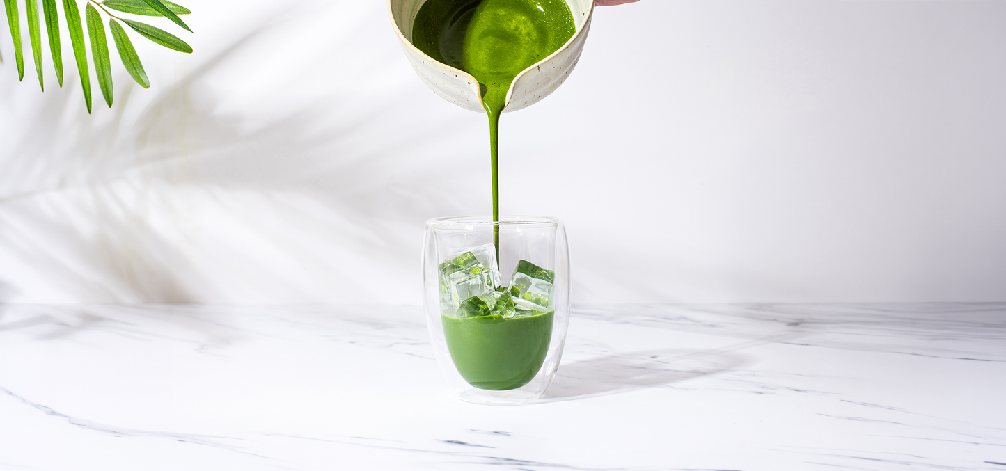 Ceremonial grade matcha being poured from a ceramic chawan into an iced matcha latte glass on a marble surface