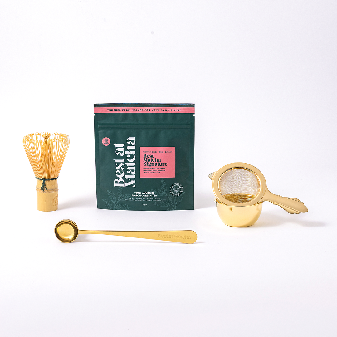Matcha tea set with matcha powder pouch, whisk, gold measuring scoop, and strainer on a white background