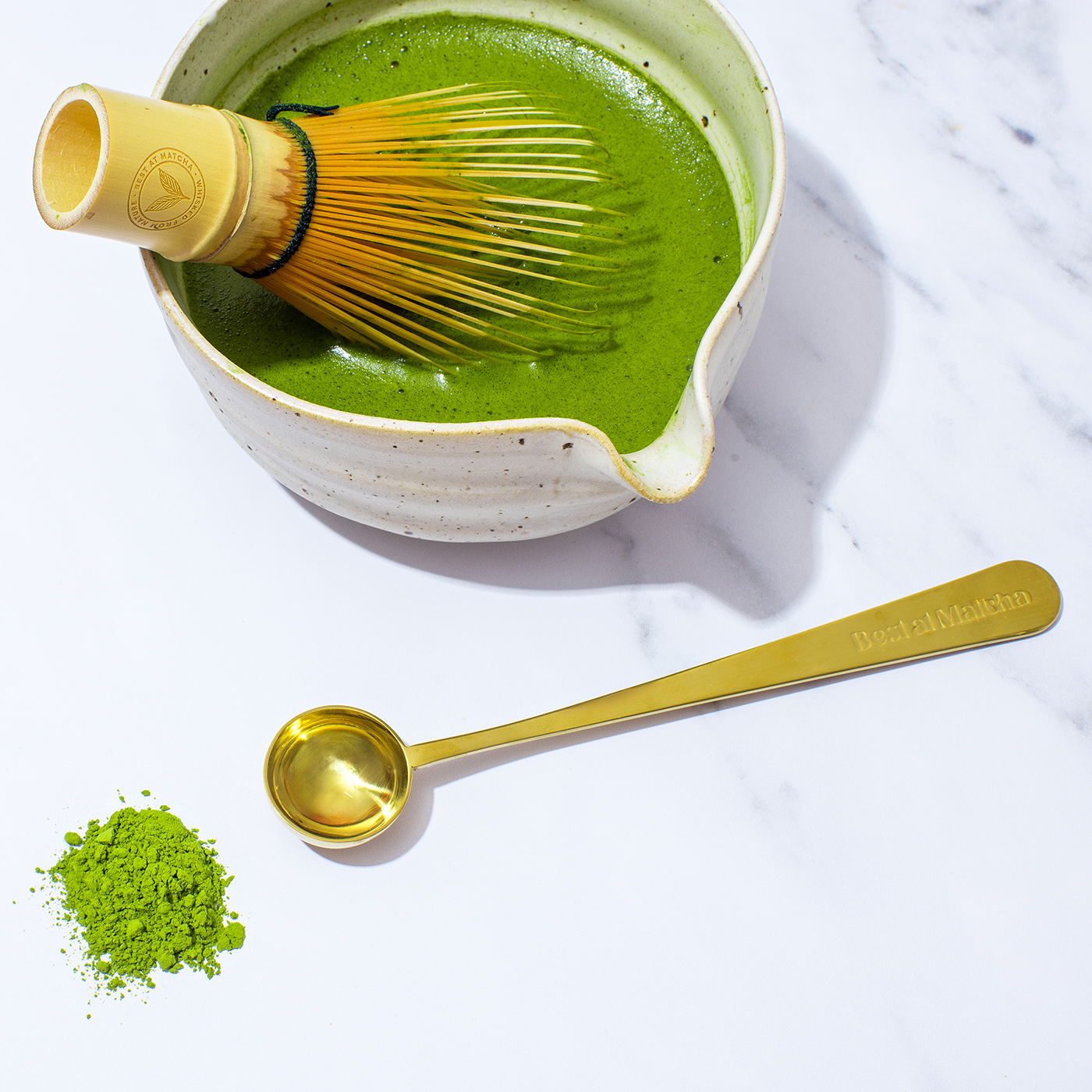 Matcha green tea in a bowl with a whisk, spoon, and matcha powder on a white background